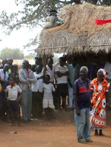 A man on stilts in the mask "Mfiti Ilauala," leaning against the roof of the house where the dead chief's body was laid.
